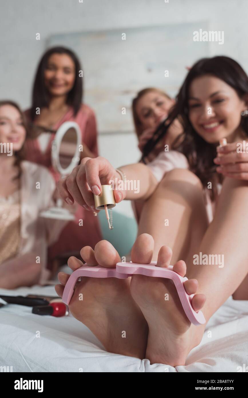 Foyer sélectif de la jeune fille brunette souriant et peindre des ongles avec vernis à ongles avec des amis multiethniques sur le lit Banque D'Images