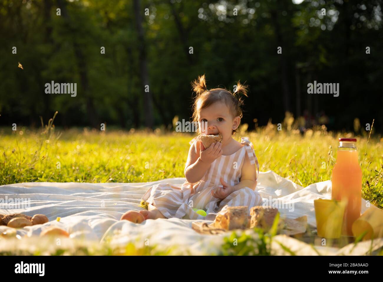 Petite fille dans une robe rayée sur un pique-nique dans un parc de la ville. Elle mange de la baguette fraîche et des fruits, boit du jus naturel. L'harmonie de l'homme et de la nature. Banque D'Images