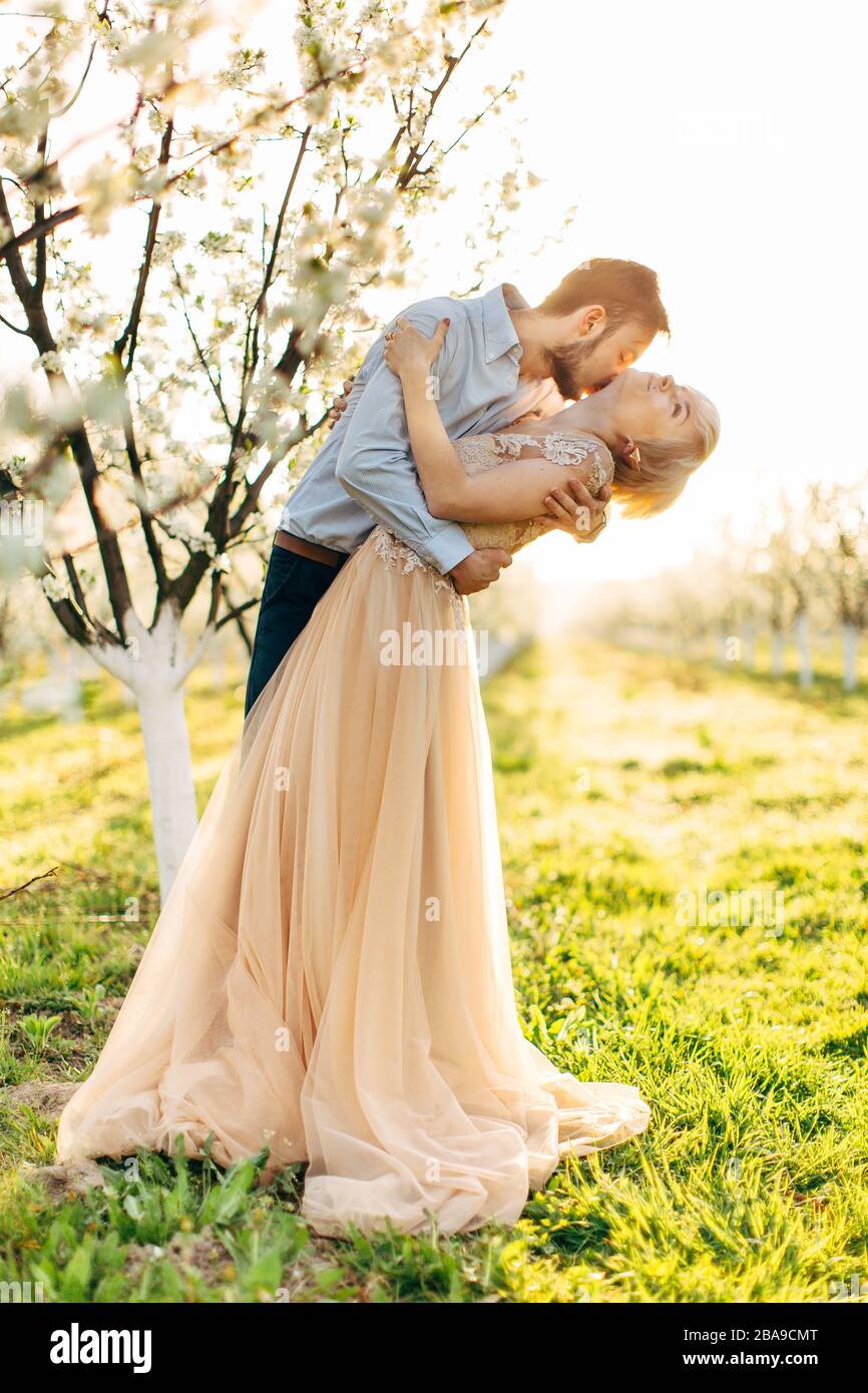 Beau couple, gai et romantique amoureux se posant dans le jardin fleuri. Bel homme embrassant le cou de sa jolie femme dans une robe élégante avec de grandes Banque D'Images