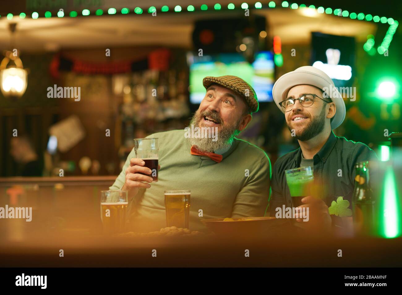 Des hommes souriants bbarbus boivent de la bière et regardent le sport tout en étant assis au bar sportif Banque D'Images