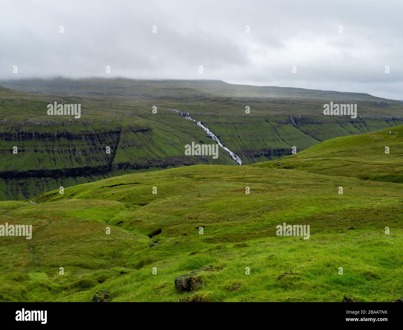 Vue typique des îles Féroé. Champs d'herbe verte, chute d'eau et nuages au-dessus des collines. Paysage brumeux. Banque D'Images