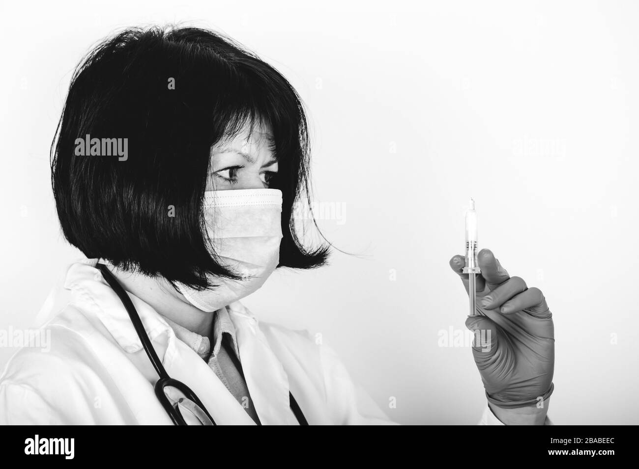 Médecin féminin en blouse de laboratoire blanche et masque médical sur fond blanc contenant seringue et médicament en ampoule. Photo en noir et blanc. Banque D'Images