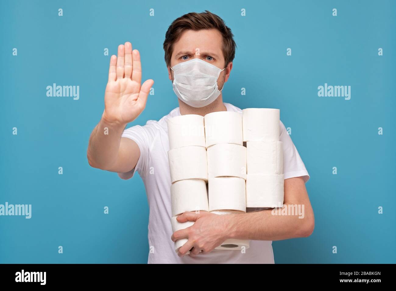 Cupidité et panique à cause du coronavirus. Homme dans le masque médical tenant beaucoup de rouleaux de papier toilette. Photo studio sur mur bleu Banque D'Images