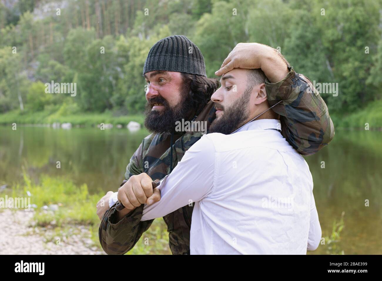 Un terroriste barbu brutal dans le camouflage garde le long couteau au cou du jeune homme terrifié dans une chemise blanche sur la rive du lac. Prise d'otages, threa terroriste Banque D'Images