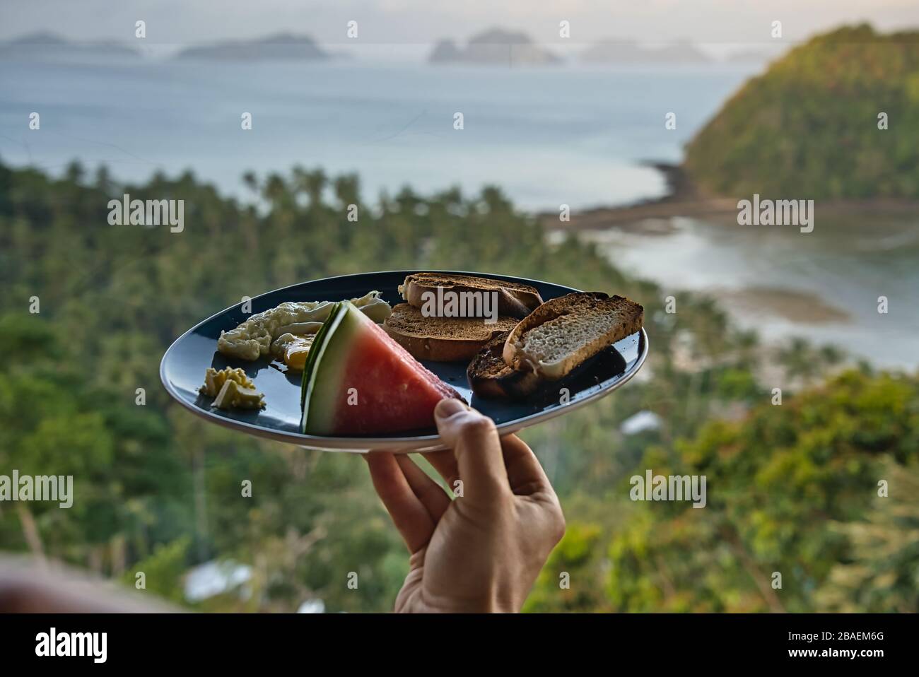 Homme transportant un petit déjeuner continental sain en face d'une plage paradisiaque surplombant El Nido, Philippines. Pastèque, œufs brouillés, pain grillé et beurre Banque D'Images