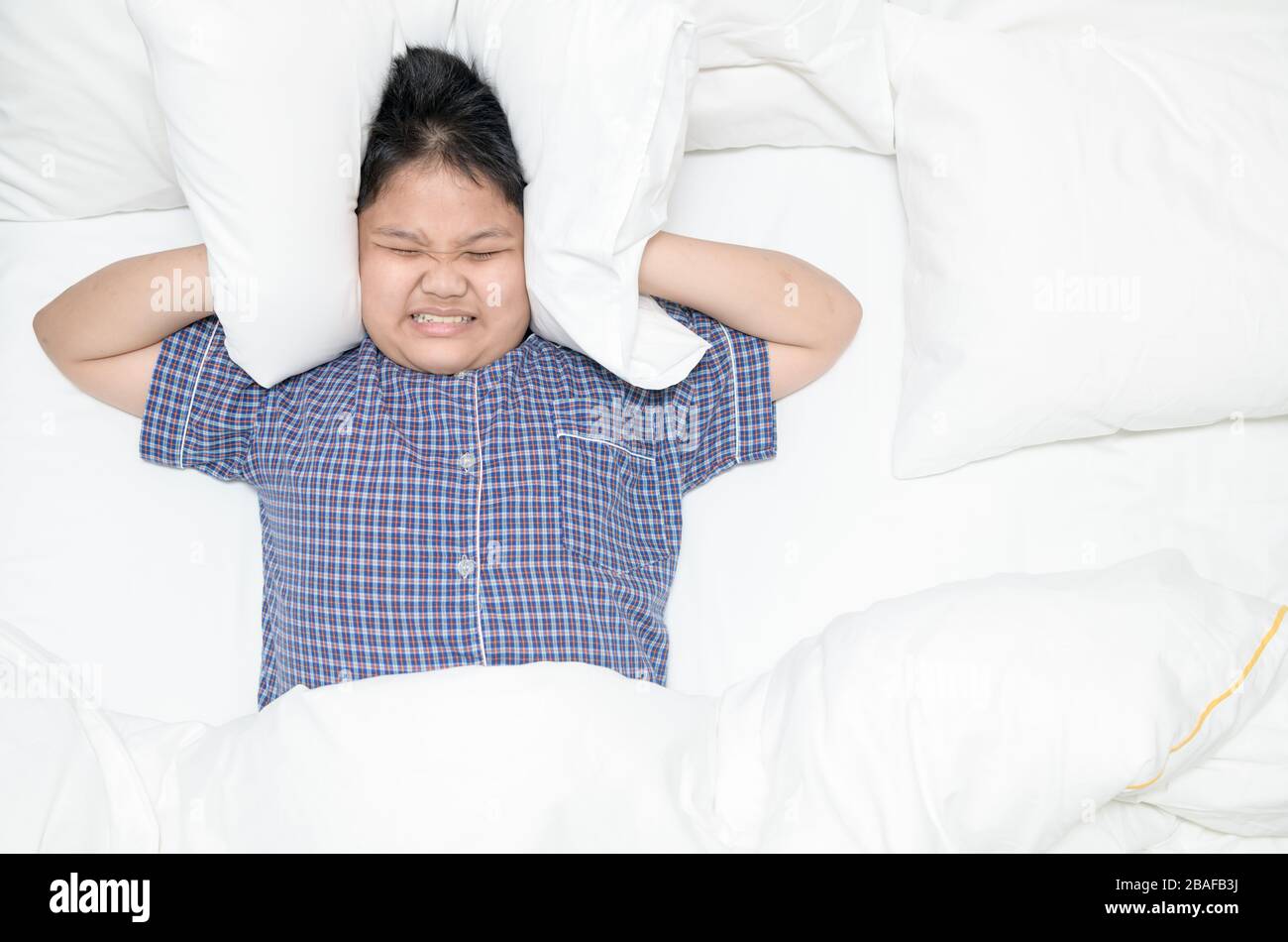 Gros garçon obèse allongé dans le lit couvrant la tête avec un oreiller parce que trop de bruit gênant. Enfant irrité souffrant de voisins bruyants, essayant de dormir Banque D'Images