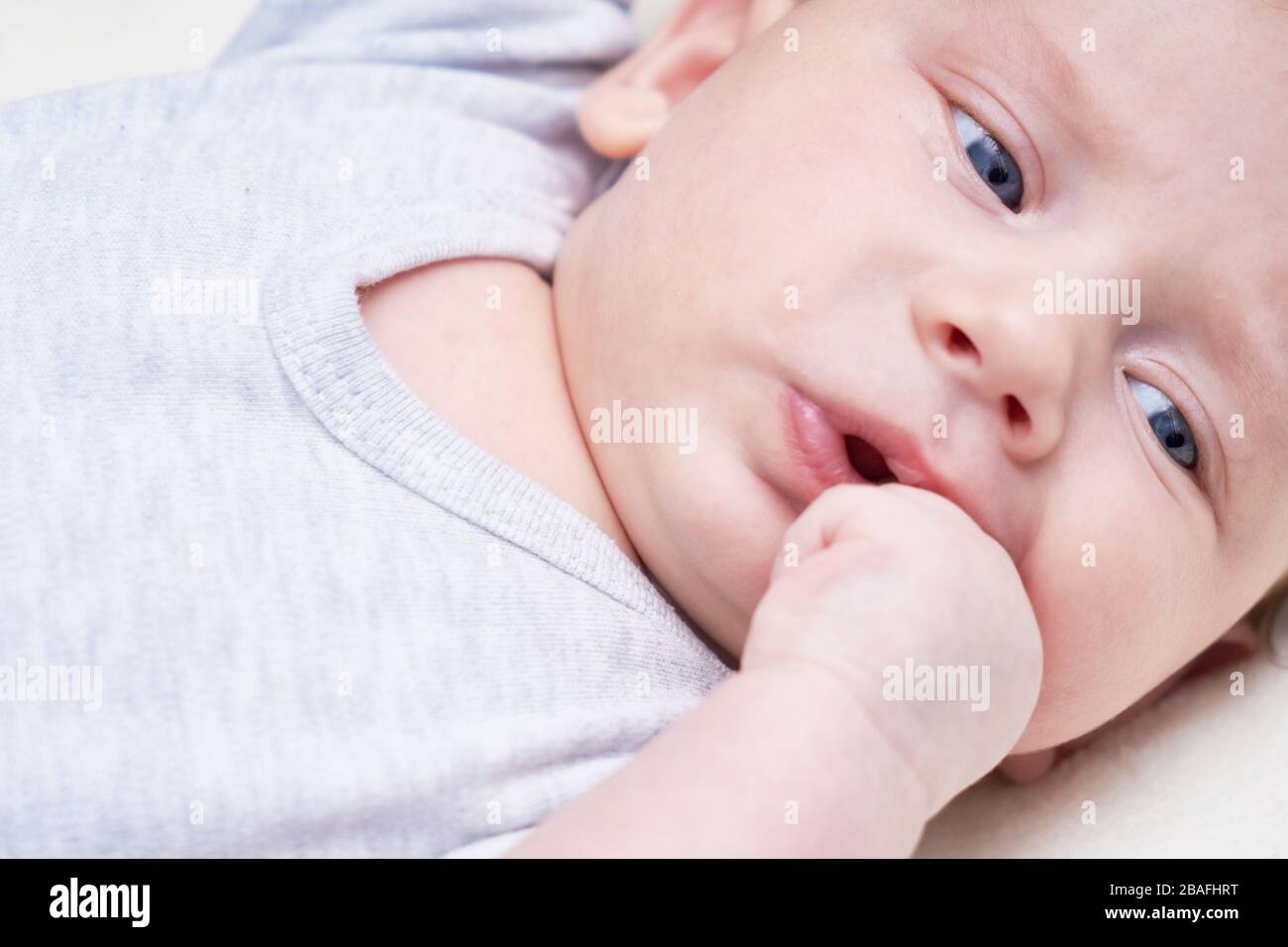 Bébé mignon avec yeux bleus suce les doigts, poing. Sur fond clair Banque D'Images