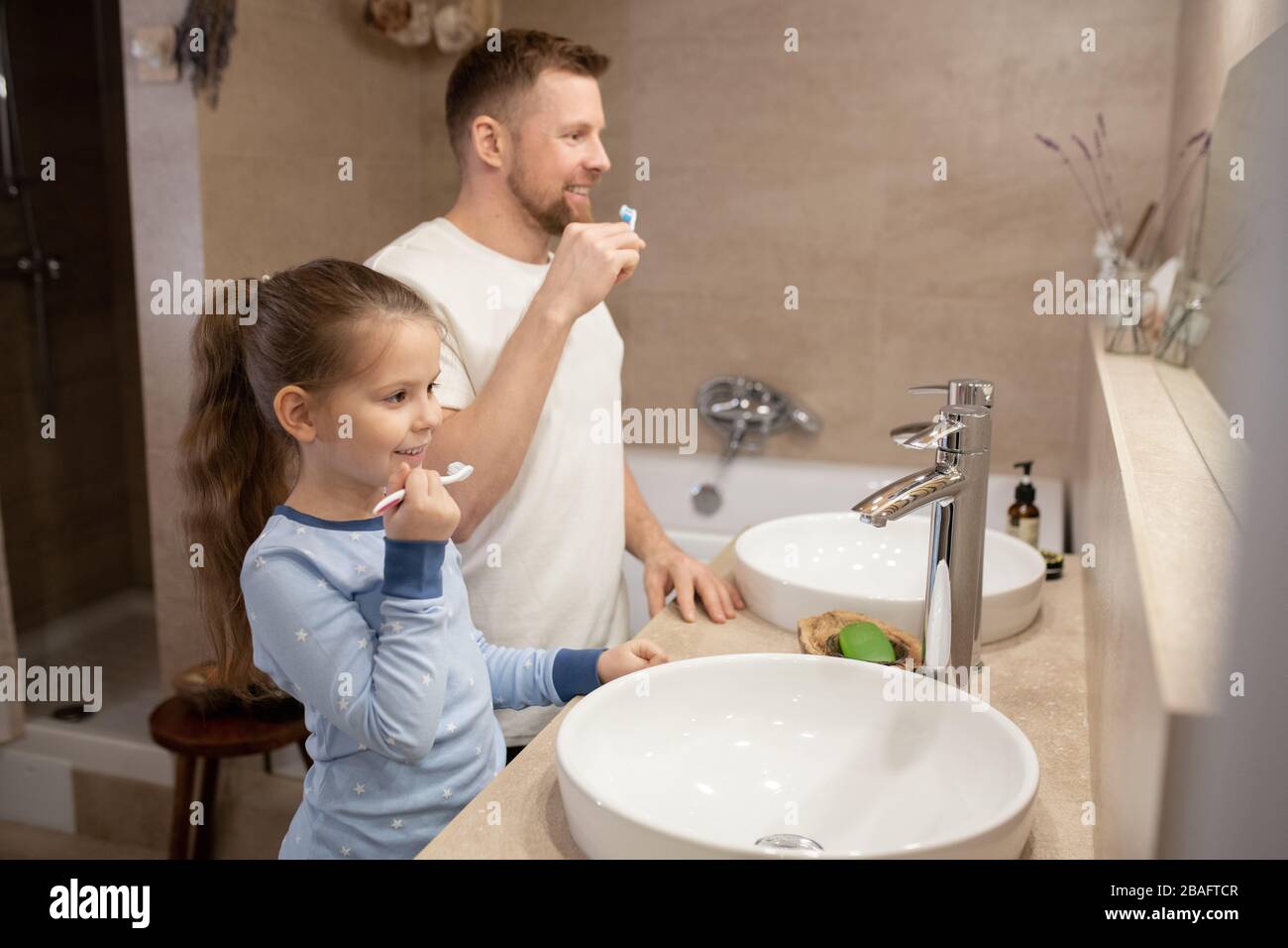 Jolie petite fille et son jeune père heureux avec brosses à dents allant brosser les dents tout en se tenant dans la salle de bains devant le miroir Banque D'Images