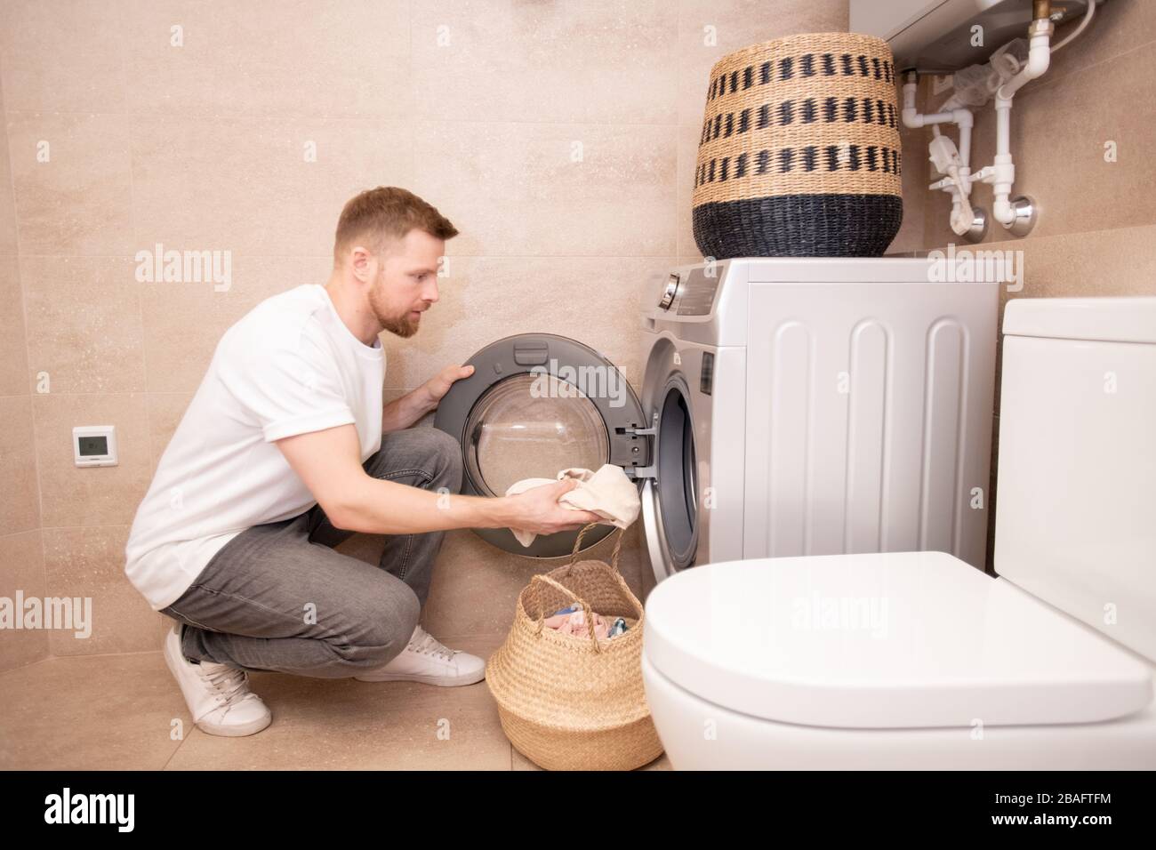 Jeune homme prenant la serviette sale du panier et les mettre dans la machine à laver tout en squatant contre le mur dans la salle de bains Banque D'Images