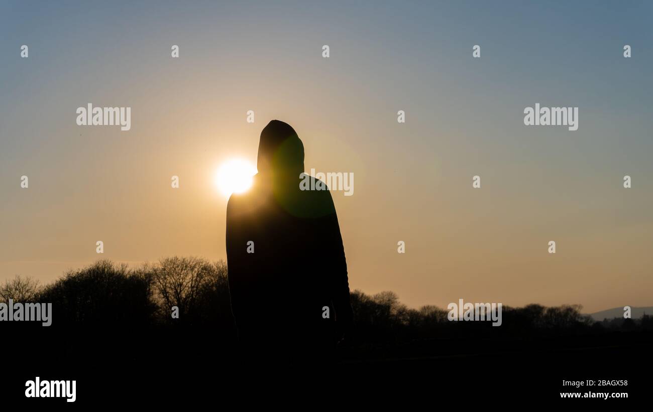 Une figure cagoule sombre et tassée de silhouetted contre le coucher du soleil. Avec lumière diffuse délibérée. Banque D'Images