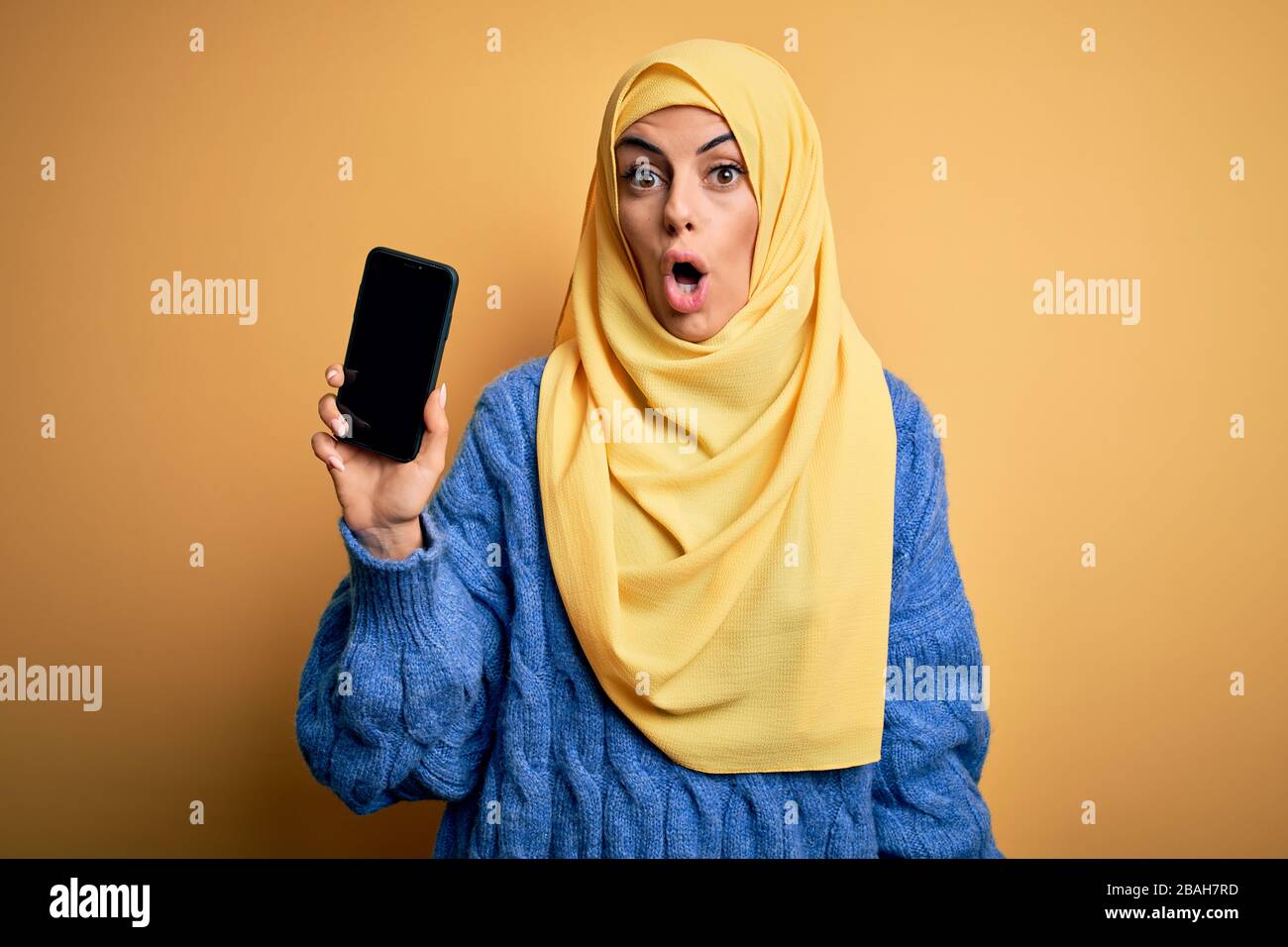 Jeune femme arabe brunette portant un hijab islamique tenant smartphone montrant écran peur en choc avec un visage surprise, peur et excité W Banque D'Images