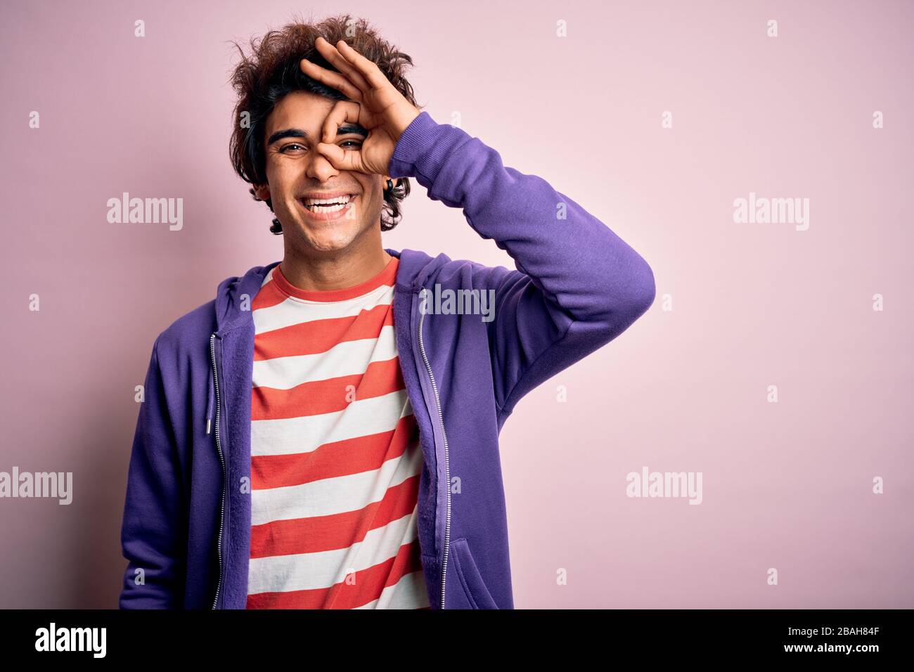 Jeune homme portant un t-shirt rayé et un sueur pourpre sur un fond rose isolé faisant un geste ok avec sourire à la main, oeil regardant à travers les doigts W Banque D'Images
