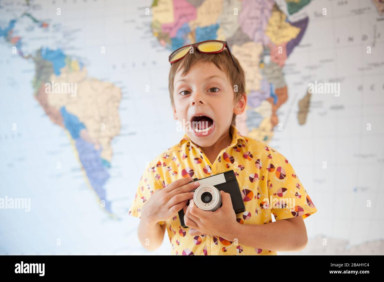 drôle petit enfant hurlant dans les lunettes de soleil et chemise jaune tenant l'appareil photo vintage sur le fond de l'homme du monde Banque D'Images