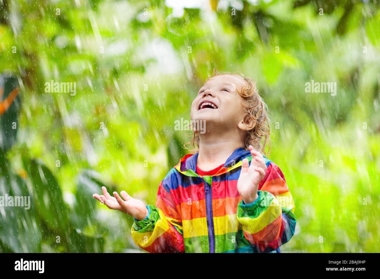 Enfant jouant sous la pluie le jour ensoleillé de l'automne. Enfant sous une forte douche portant une veste imperméable arc-en-ciel. Petit garçon dans le parc des pluies. Activité extérieure de l'automne Banque D'Images