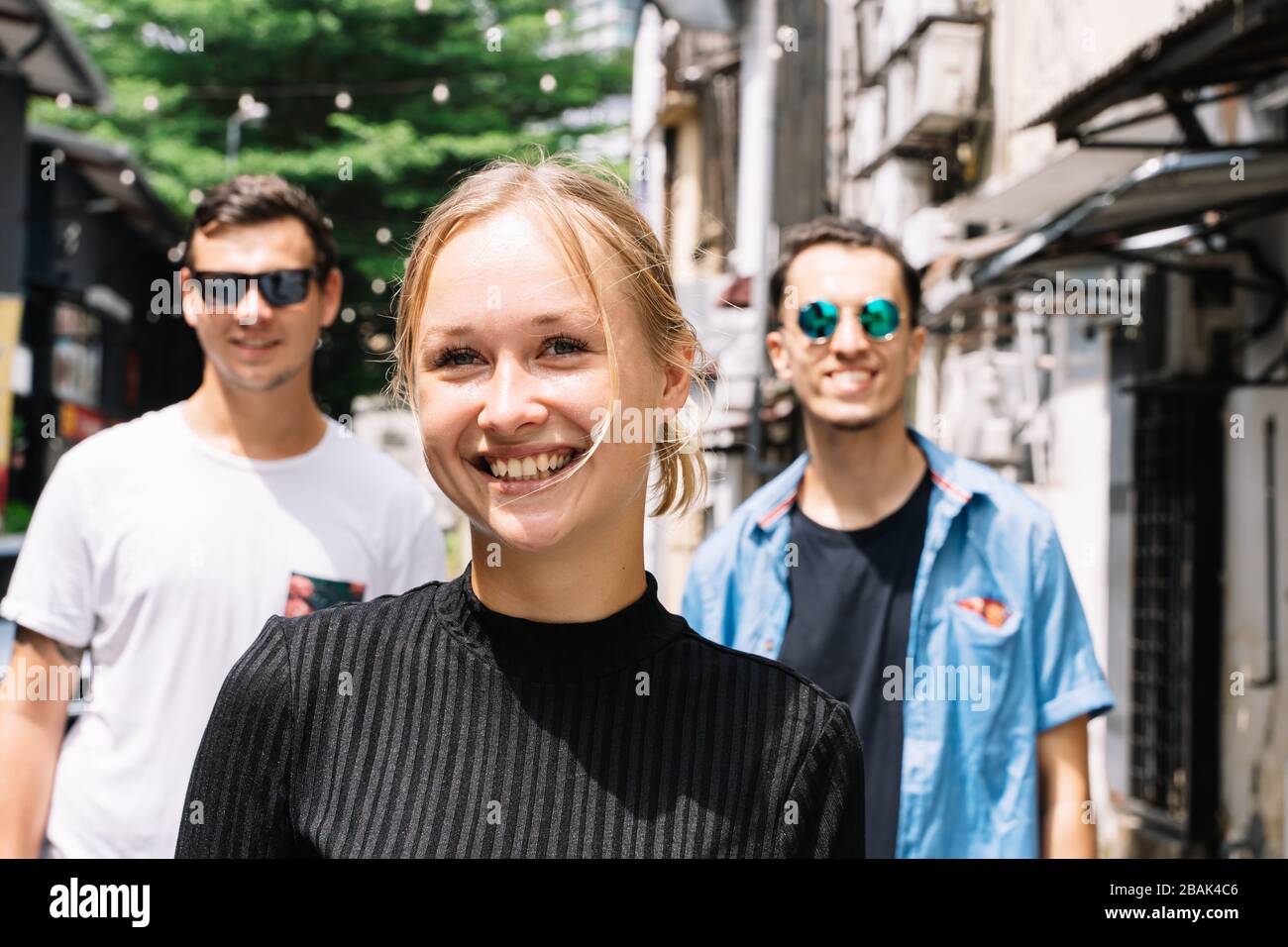 Blonde fille souriant devant deux amis dans une rue avec des arbres et des lumières accrochées dans une ville Banque D'Images