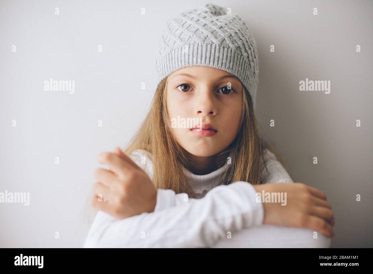 jolie petite fille caucasienne avec de longs cheveux portant un pull et un chapeau de laine assis sur un mur brillant penchant regardant dans l'appareil photo Banque D'Images