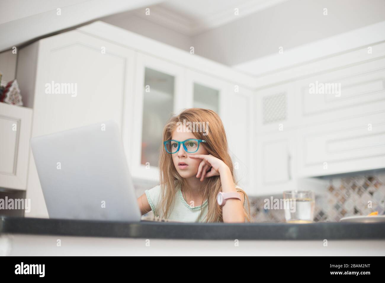 jolie petite fille dans des verres assis à la cuisine avec ordinateur portable en ligne surfer sur internet Banque D'Images