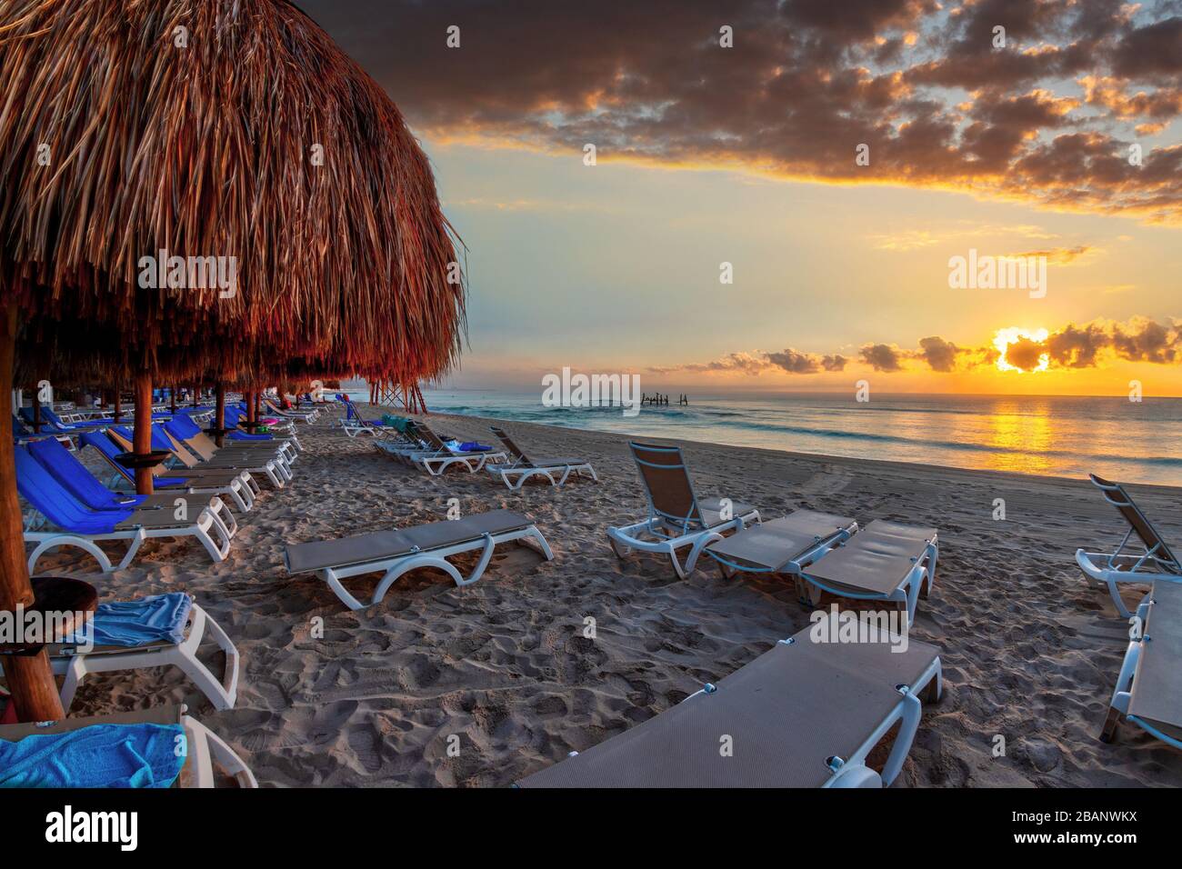 Lever du soleil doré sur des rangées de chaises longues et de parasols de palmiers lors de vacances sur une plage de sable des Caraïbes à Riviera Maya à Cancun, au Mexique. Banque D'Images