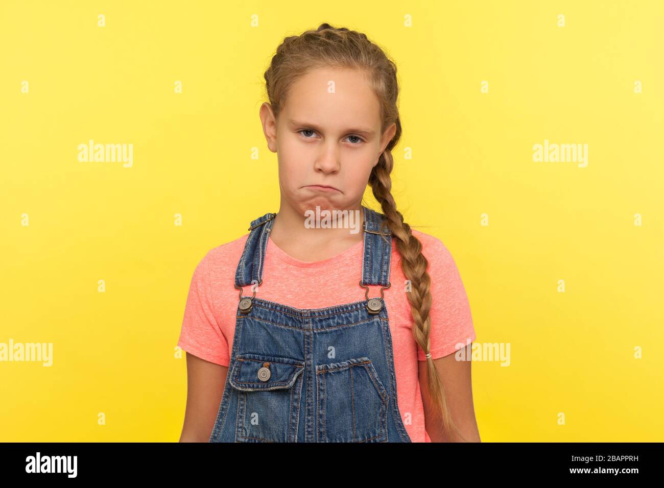 Portrait d'une petite fille mécontente et drôle et frustrée, avec une tresse dans des salopettes en denim, regardant un appareil photo avec une expression de visage mécontent et comique. Intérieur Banque D'Images