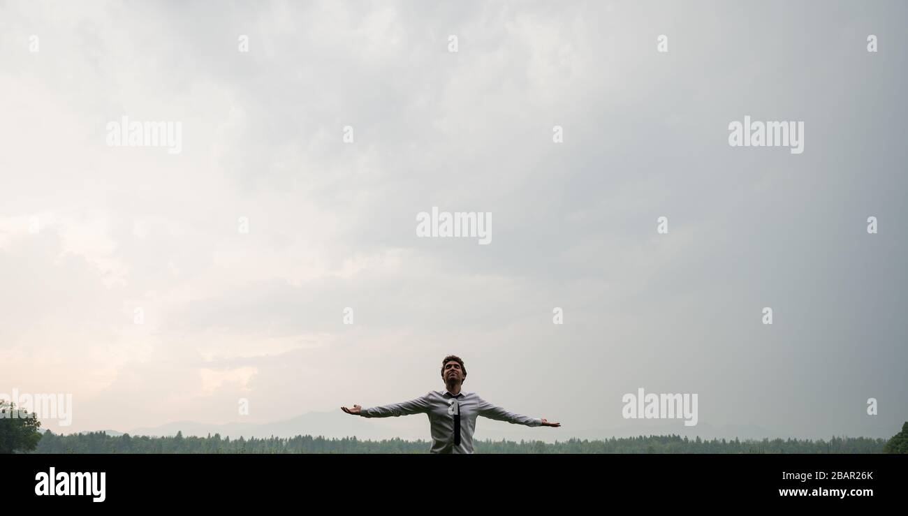 Jeune homme d'affaires debout avec ses bras se propagent sous un ciel gris nuageux, se rechargeant après une longue journée stressante. Banque D'Images