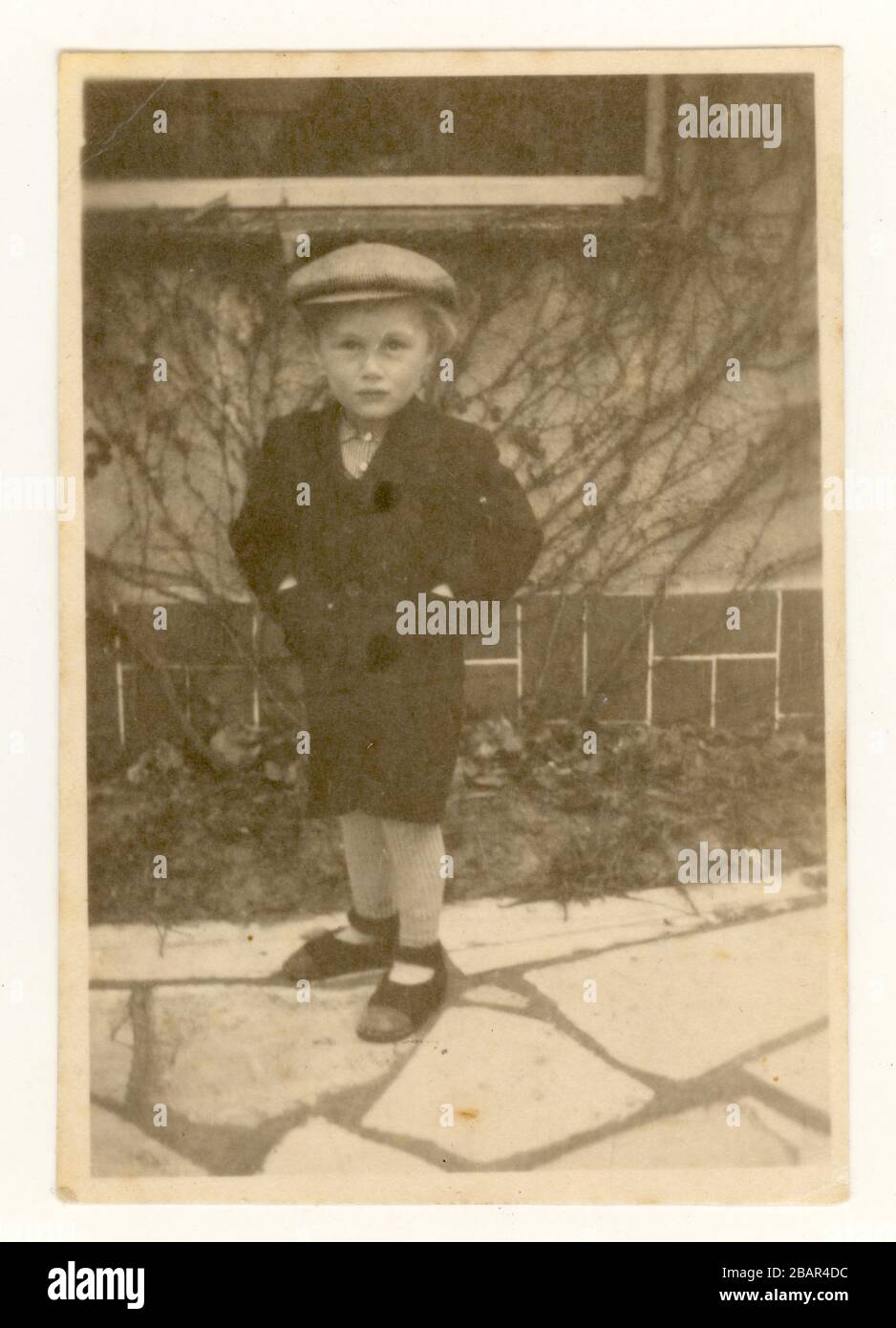 Début du XXe siècle, la photo délavée d'un jeune garçon à l'extérieur sur un patio portant un chapeau plat, l'utilisation d'un revêtement fou dans le jardin date la photo vers les années 1930, au Royaume-Uni Banque D'Images