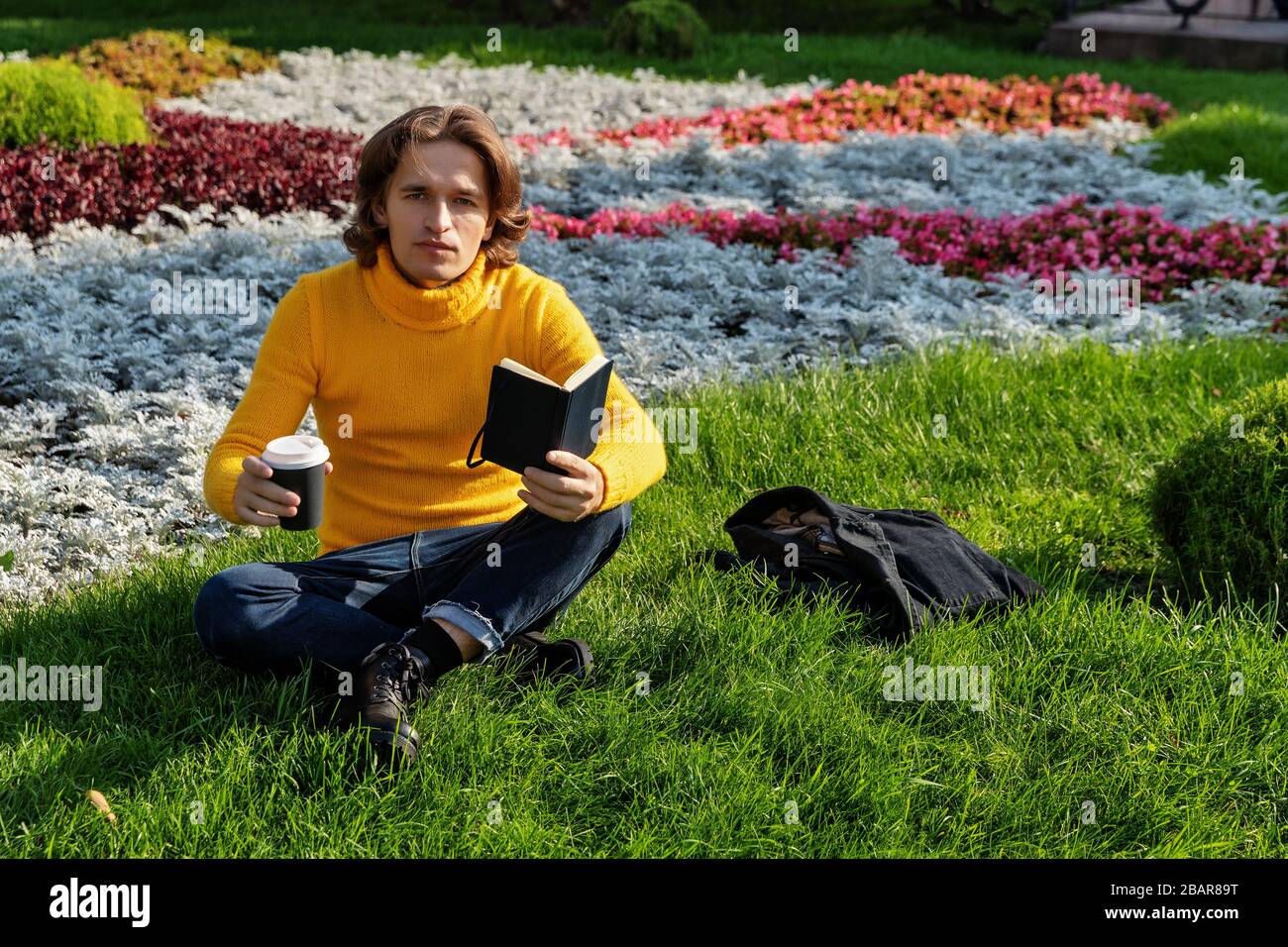 Le jeune homme boit du café et lit le livre dans le parc, il sourit, a un repos, est vêtu d'un pull jaune, des fleurs et de l'herbe sur fond Banque D'Images