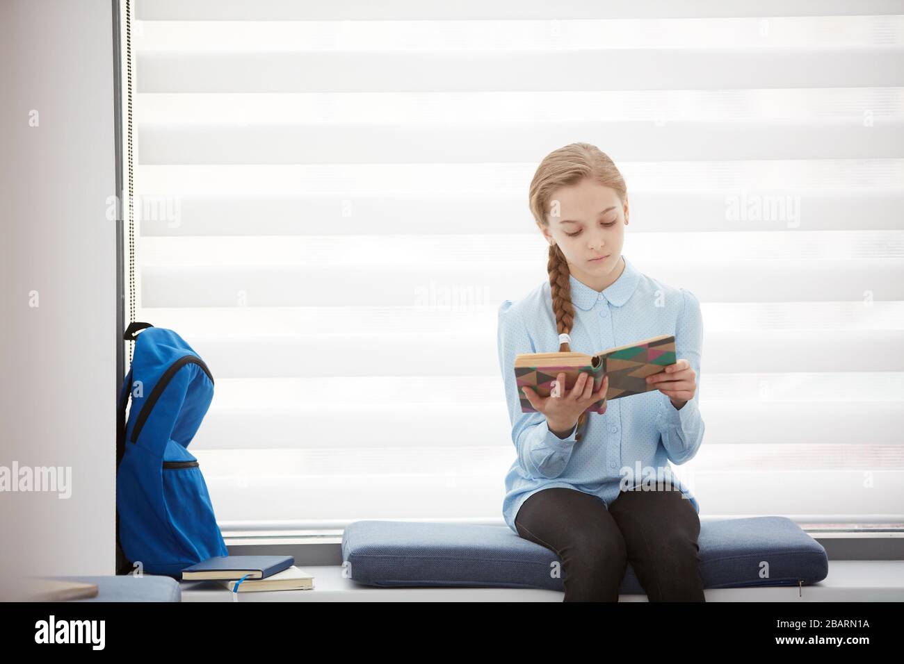 Portrait de l'adorable livre de lecture de fille d'école tout en étant assis sur la fenêtre dans la salle de classe d'école, espace de copie Banque D'Images