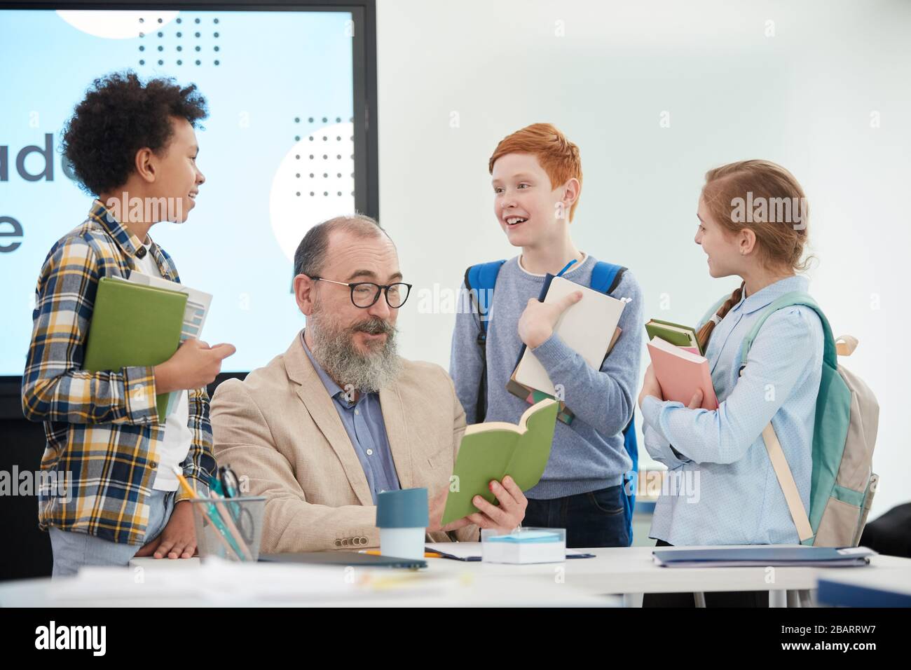 Groupe multiethnique d'enfants gaies se tenant autour de l'enseignant senior barbu dans la classe scolaire Banque D'Images