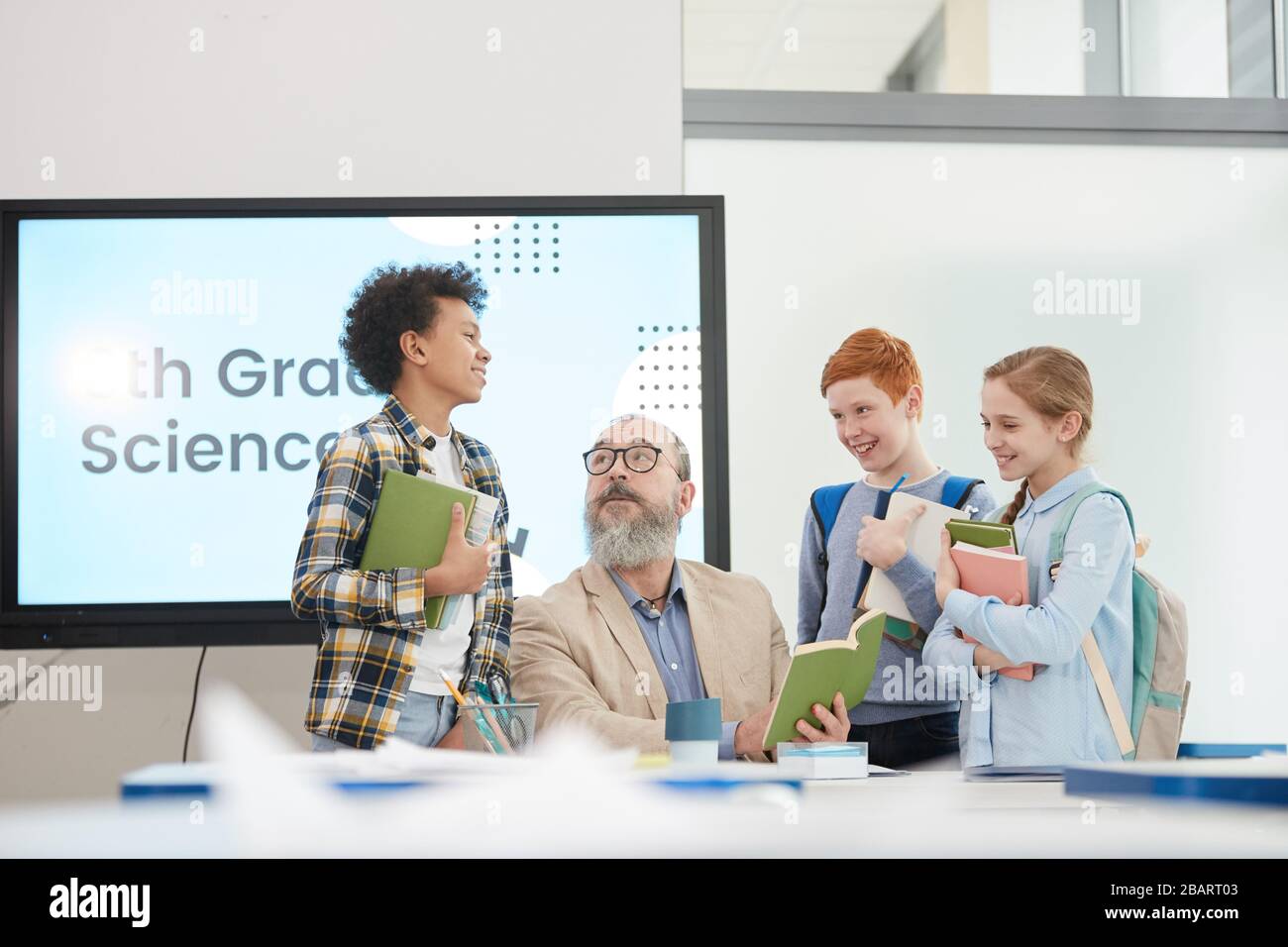 Groupe multiethnique d'enfants gaies se tenant autour de l'enseignant senior barbu dans la classe scolaire, espace de copie Banque D'Images