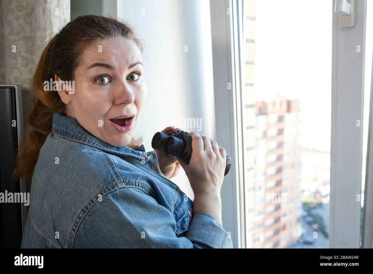 Une femme adulte choquée après avoir espionne la fenêtre avec des jumelles, une chambre domestique Banque D'Images