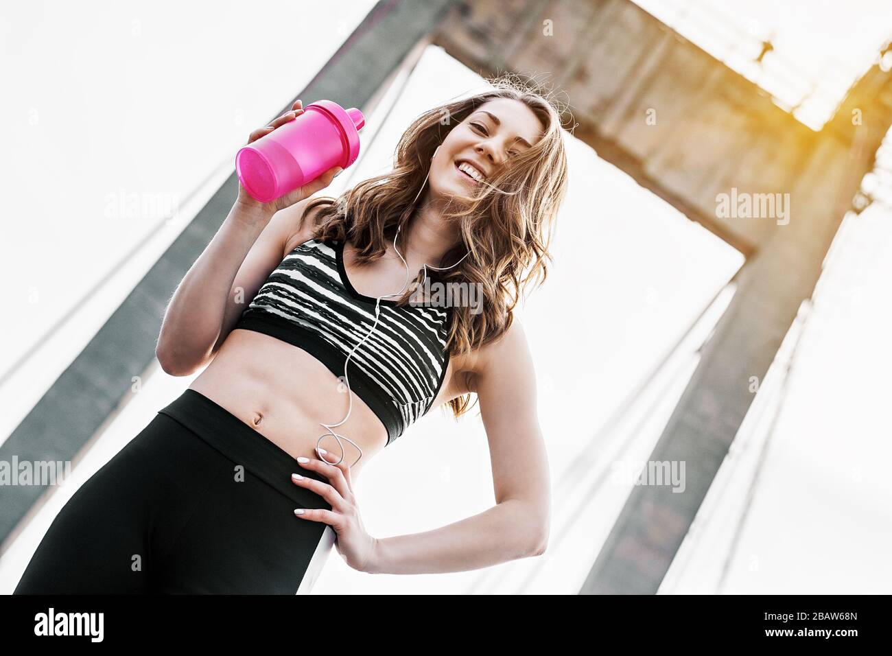 Bonne fille de sport tenant shaker bouteille d'eau dans la main et souriant tout en faisant de l'exercice sur le pont. Une jeune femme sportive séduisante à l'écoute Banque D'Images