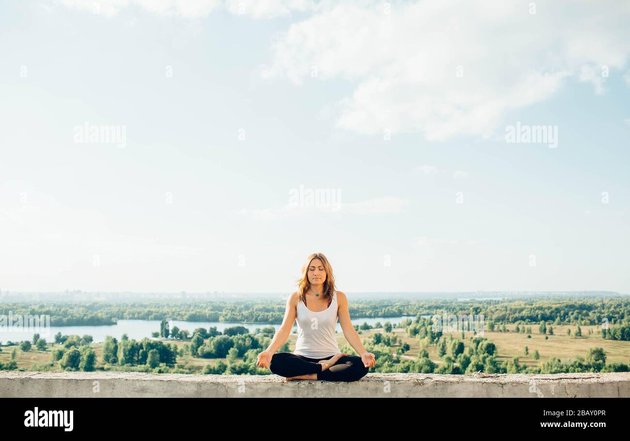 La jeune femme pratique le yoga à l'extérieur. Calme paisible fille assise sur parapet en position lotus. Ses mains baissent aux genoux. Arbres rivière et ciel sur Banque D'Images