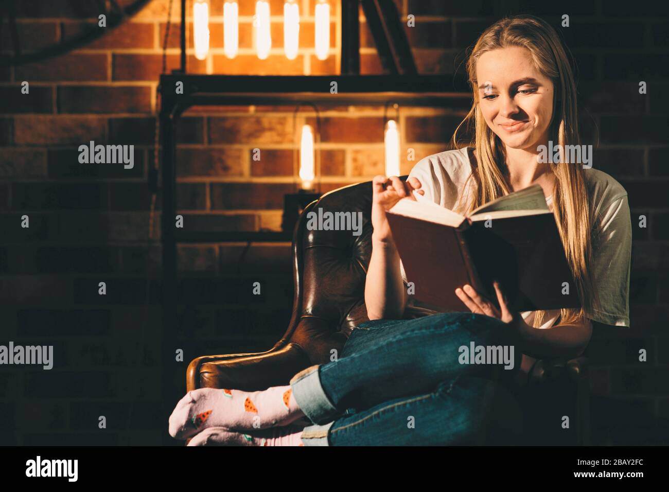 Fille dans une chambre sombre confortable lecture d'un livre avec des lunettes. Lumière blonde et chaude Banque D'Images