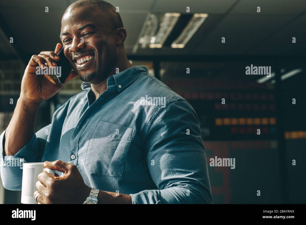 Un jeune homme souriant avec une tasse de café en main parlant sur le téléphone portable dans un bureau moderne. Un homme d'affaires heureux qui appelle au bureau. Banque D'Images