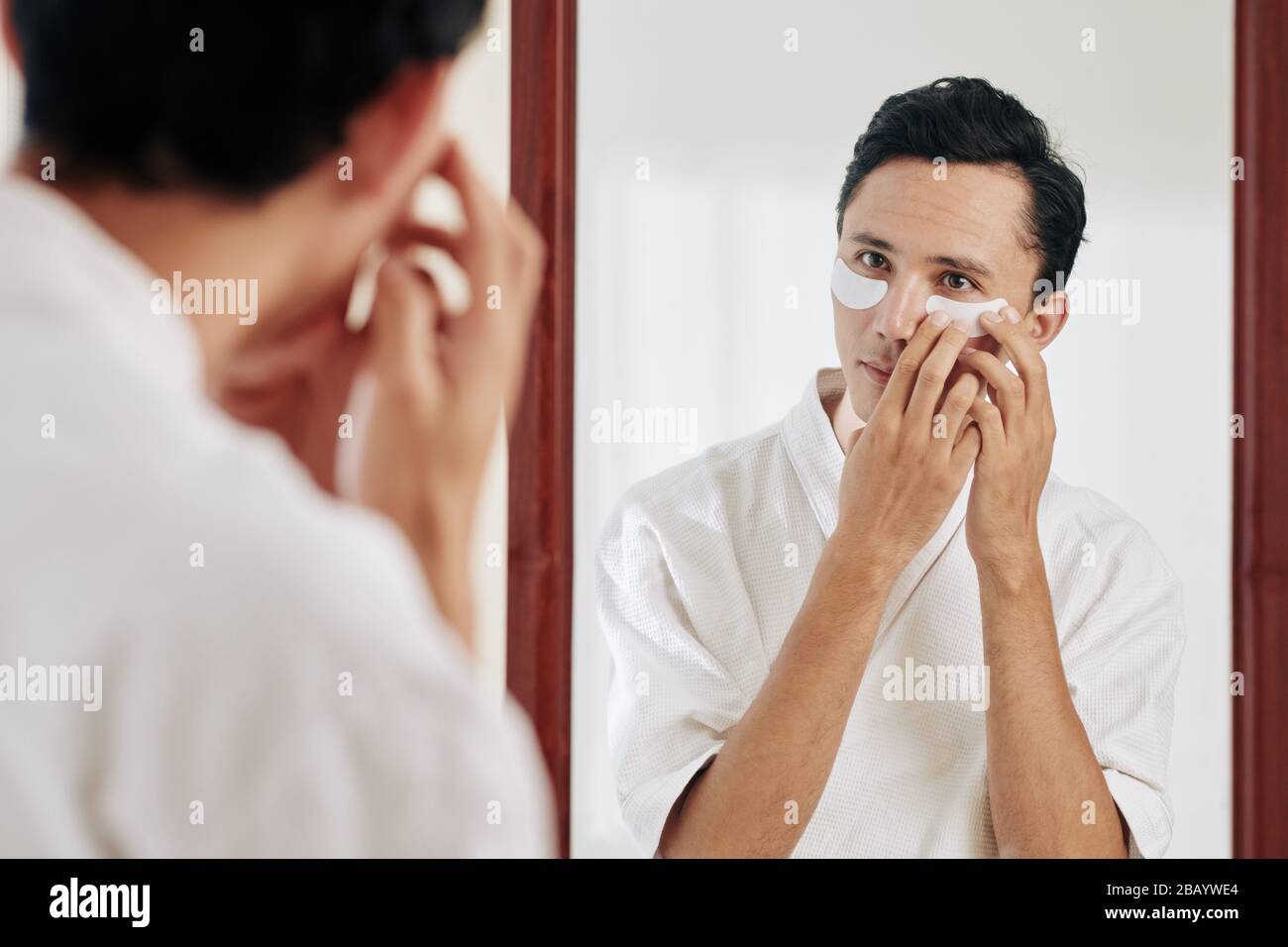 Jeune homme de course mixte debout devant le miroir et appliquant des taches sous les yeux après la douche du matin Banque D'Images