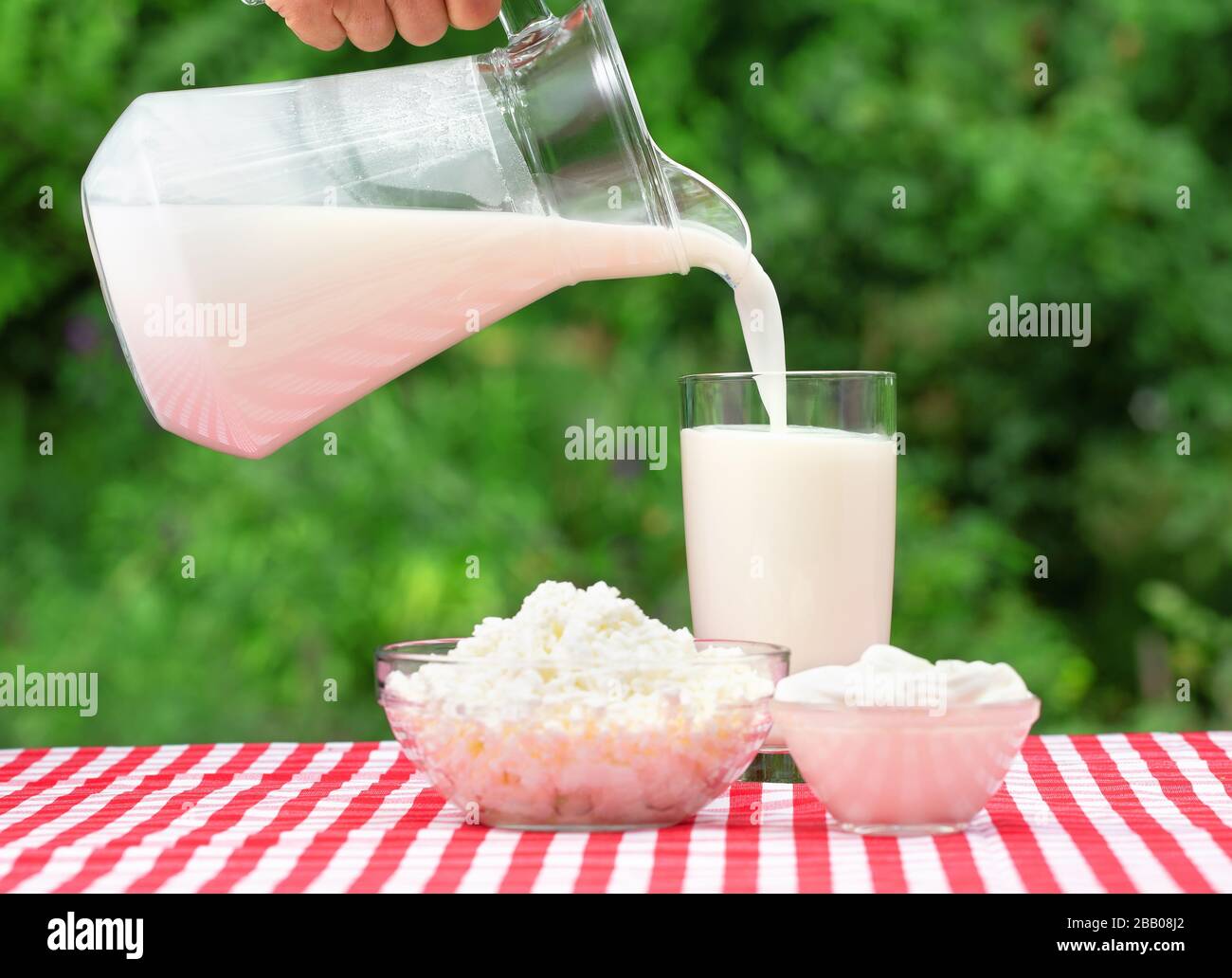 Du lait qui coule d'un pichet dans un verre. Sur la table couverte d'une nappe à damier rouge sont des produits laitiers. Fond vert naturel Banque D'Images