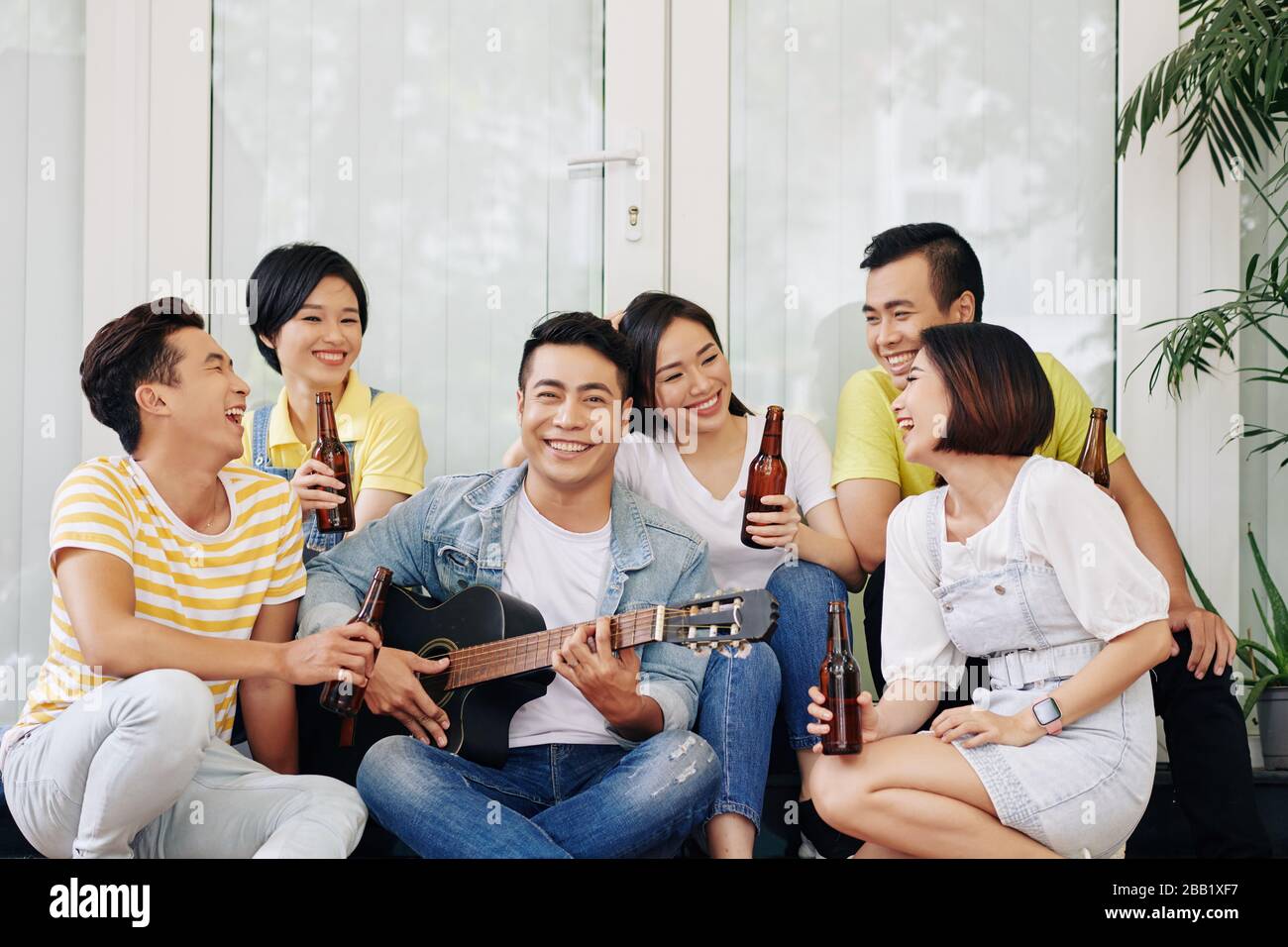Joyeux beau jeune homme vietnamien jouant de la guitare pour ses amis buvant et parler à la fête de la maison Banque D'Images