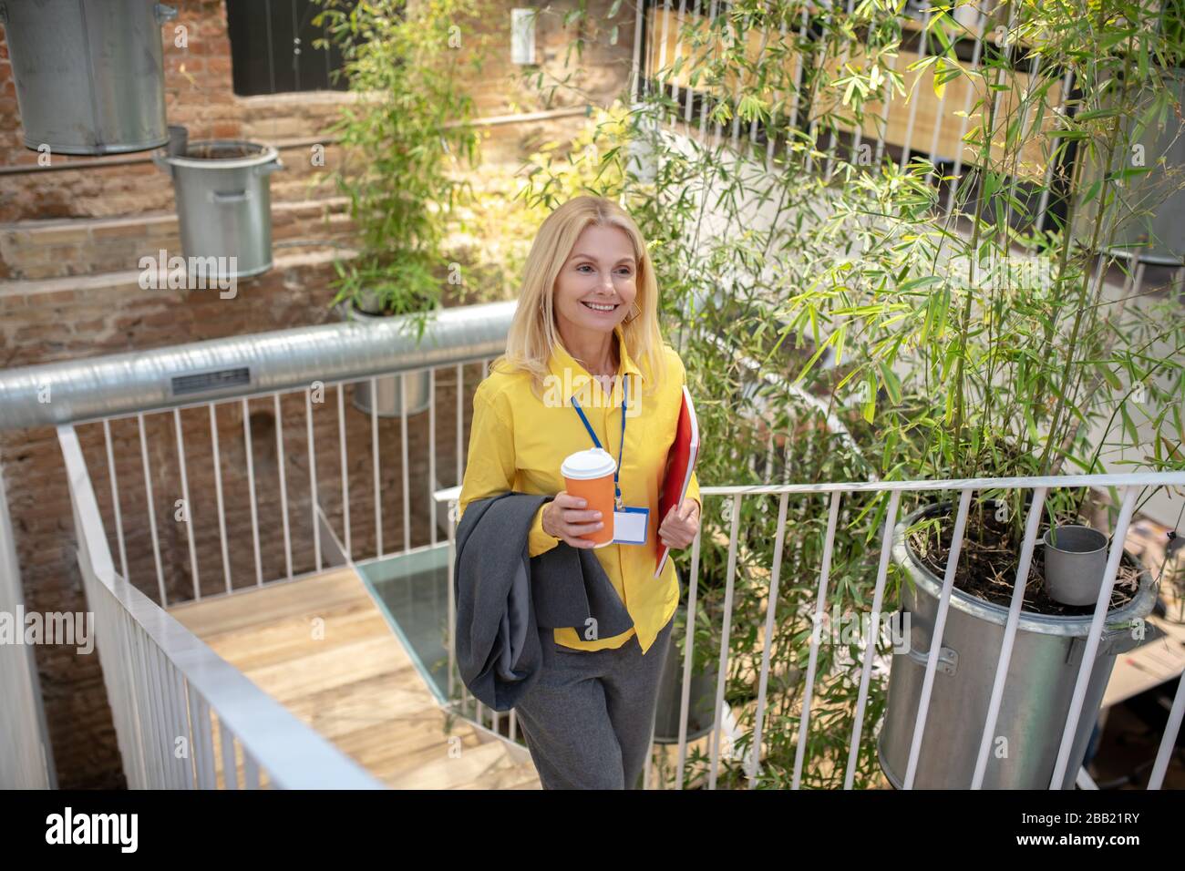 Blonde femme monter des escaliers, tenant une tasse de café et de veste, souriant Banque D'Images