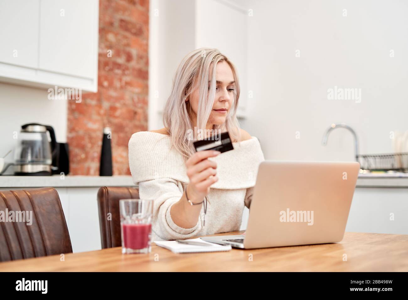 Une femme blanche avec des cheveux blonds est assise sur une table et regarde un ordinateur portable tout en tenant une carte bancaire Banque D'Images