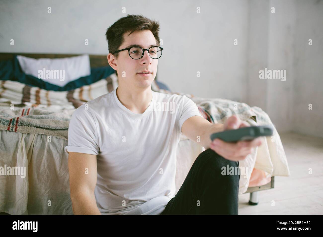 Guy utilisant la télécommande tout en étant assis sur le lit à la maison. Joyeux jeune homme qui regarde la télévision . Émotions humaines sur son visage. Banque D'Images