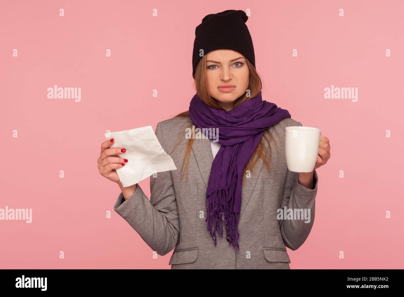 Portrait d'une femme malade irritée de la grippe dans un chapeau chaud et un foulard tenant des tissus et une tasse de boisson chaude, regardant avec une expression agacée, traitant la grippe Banque D'Images