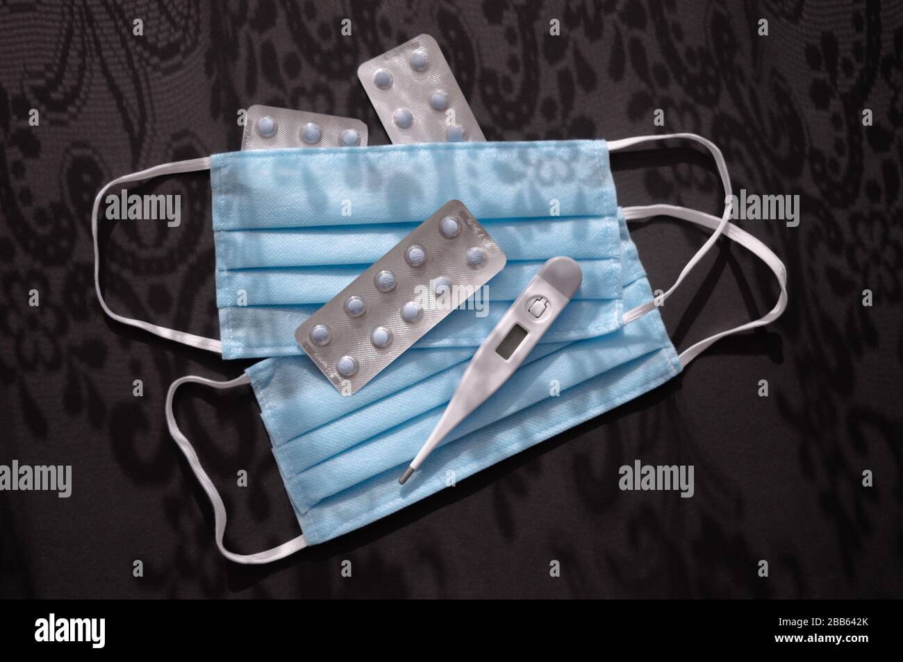 Comprimés, thermomètre et deux masques médicaux jetables bleus sur le fond sombre avec des ornements d'ombres contre le virus covid-2019. Soins de santé Banque D'Images