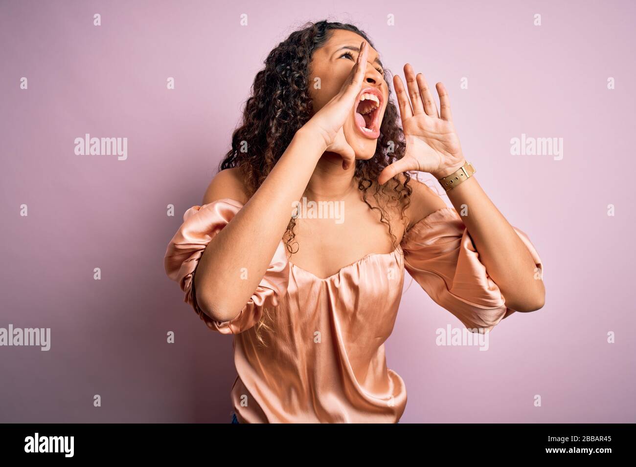 Jeune femme belle avec des cheveux bouclés portant un t-shirt décontracté debout sur fond rose criant en colère avec les mains sur la bouche Banque D'Images