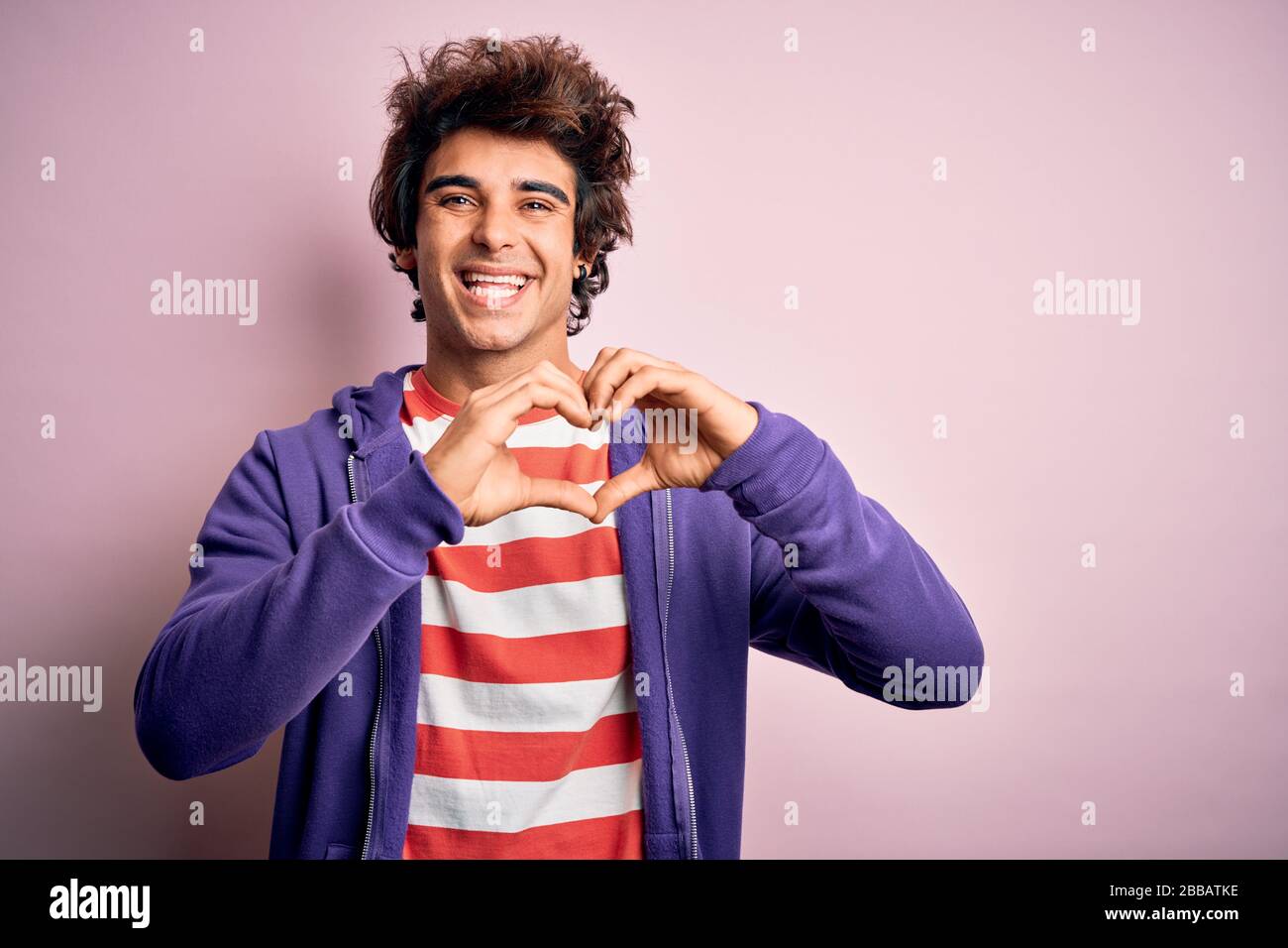 Jeune homme portant un t-shirt rayé et un sueur violet sur un arrière-plan rose isolé souriant en amour montrant le symbole du coeur et la forme avec les mains. Romant Banque D'Images