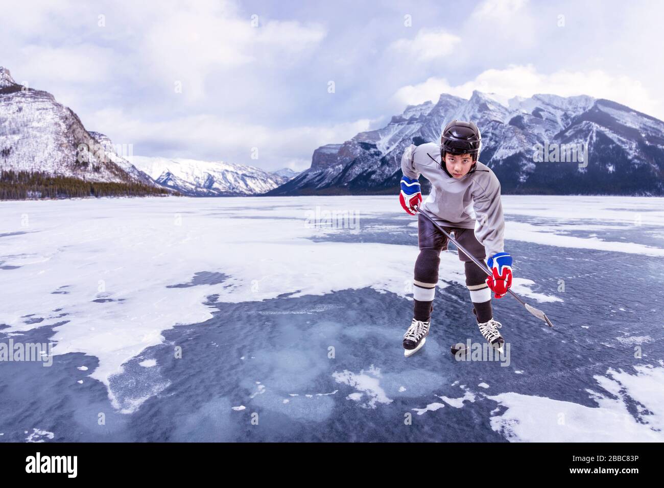 Un joueur de hockey sur glace junior portant un casque, un maillot et tenant un bâton de hockey sur palet dans une action extérieure entourée de montagnes dans les Rocheuses canadiennes. Banque D'Images