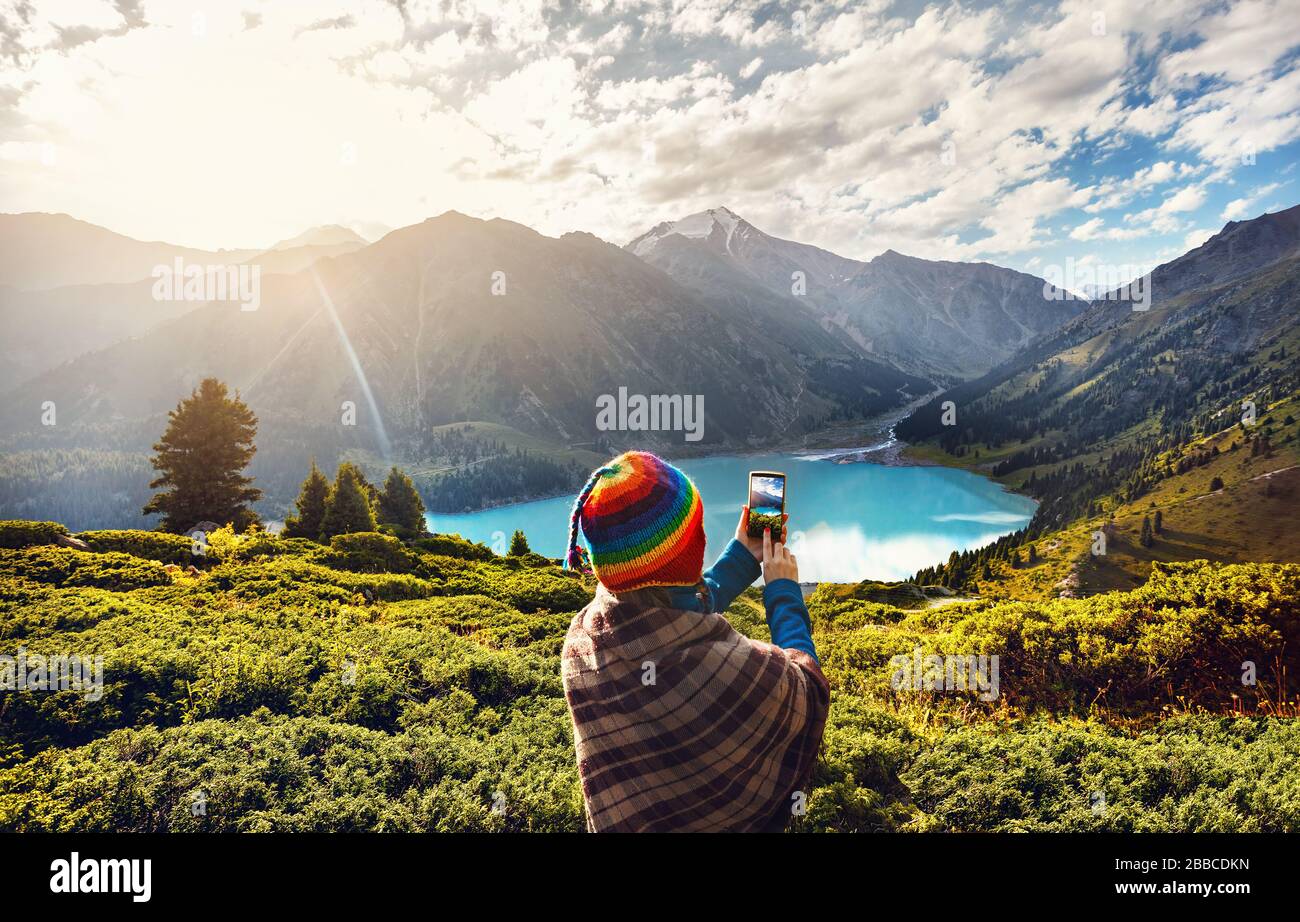 Arc-en-ciel en femme touristiques hat et marron poncho en tenant avec son smartphone photo dans le magnifique lac de montagne au Kazakhstan Banque D'Images