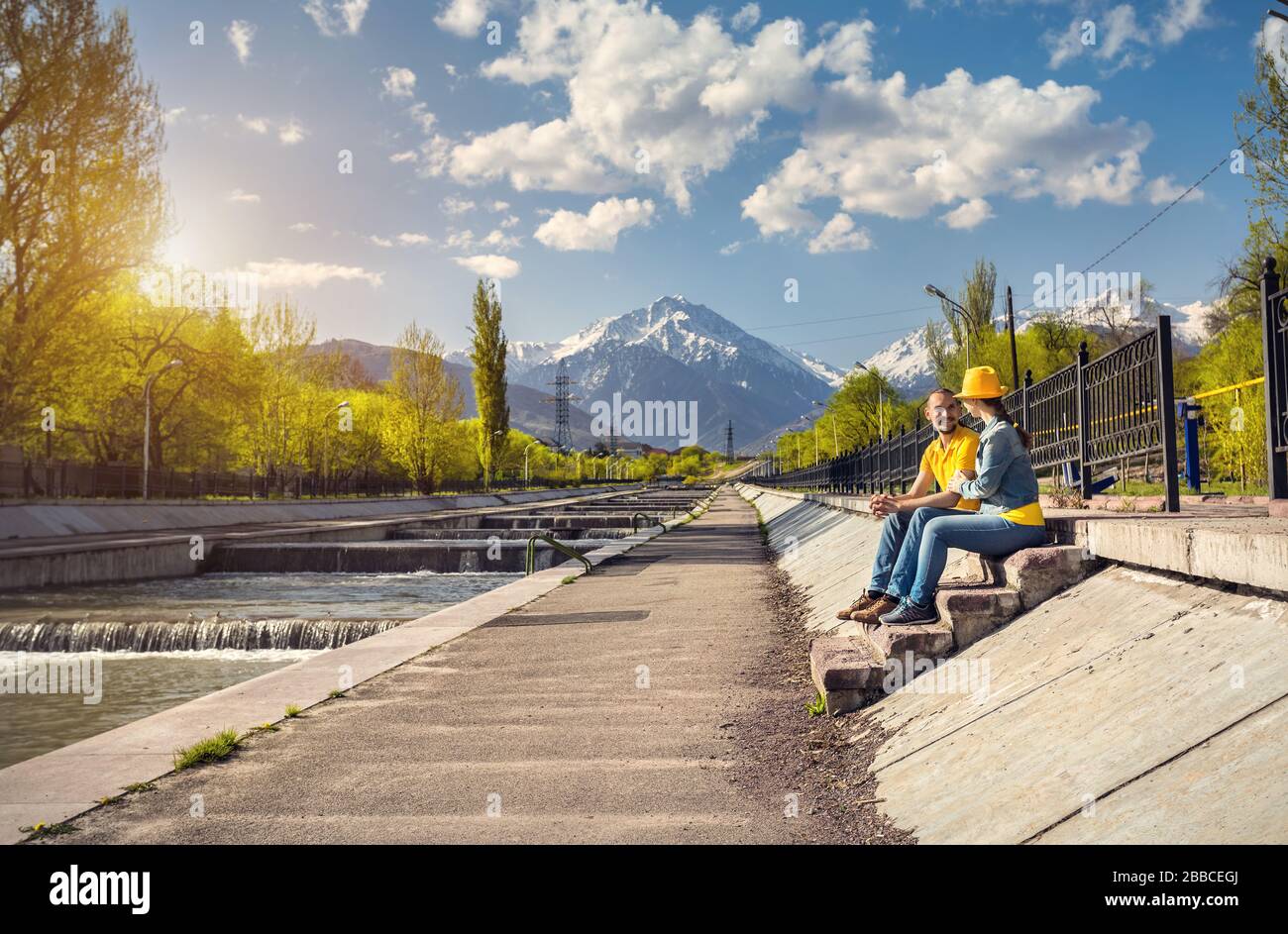 Heureux couple en costumes bleu et jaune assis sur le escaliers près de la rivière et les montagnes enneigées en arrière-plan Almaty, Kazakhstan Banque D'Images