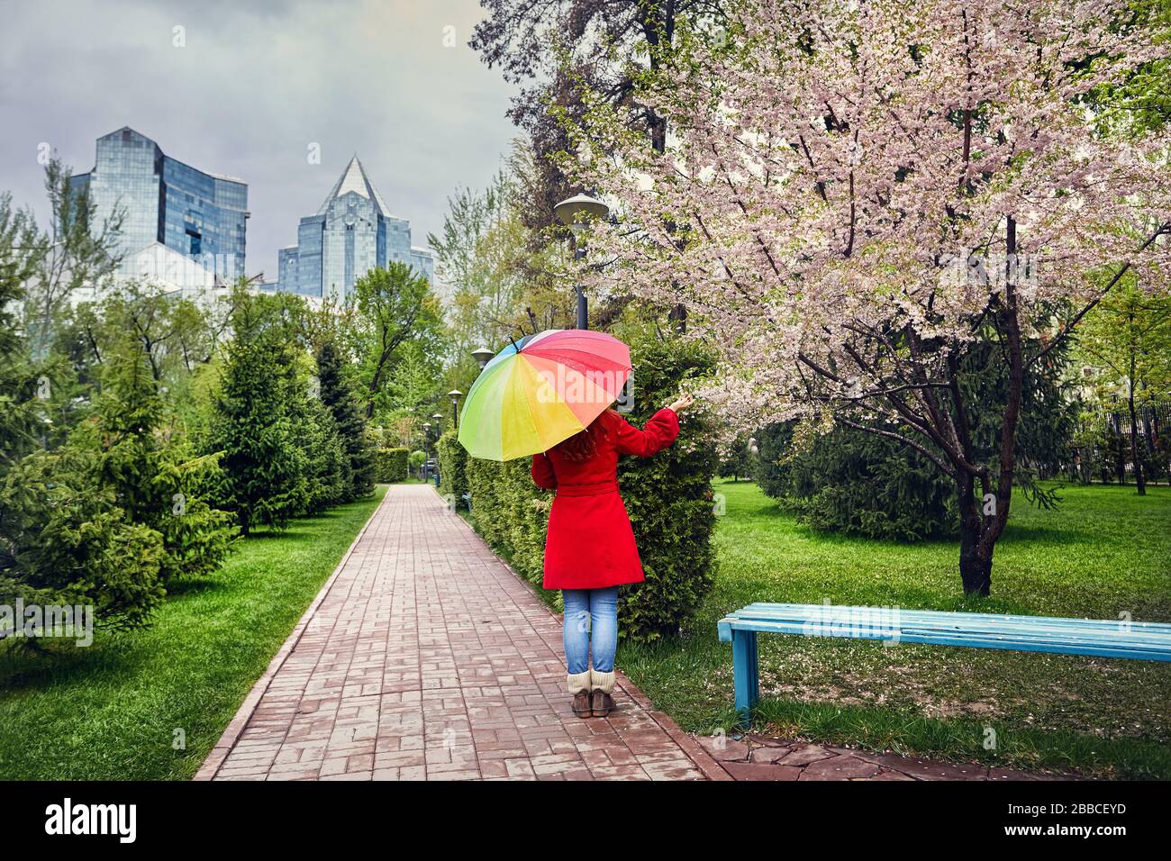 Femme en robe rouge avec parapluie arc-en-marche dans la rue, dans le parc avec ciel couvert et fleurs de cerisier Banque D'Images