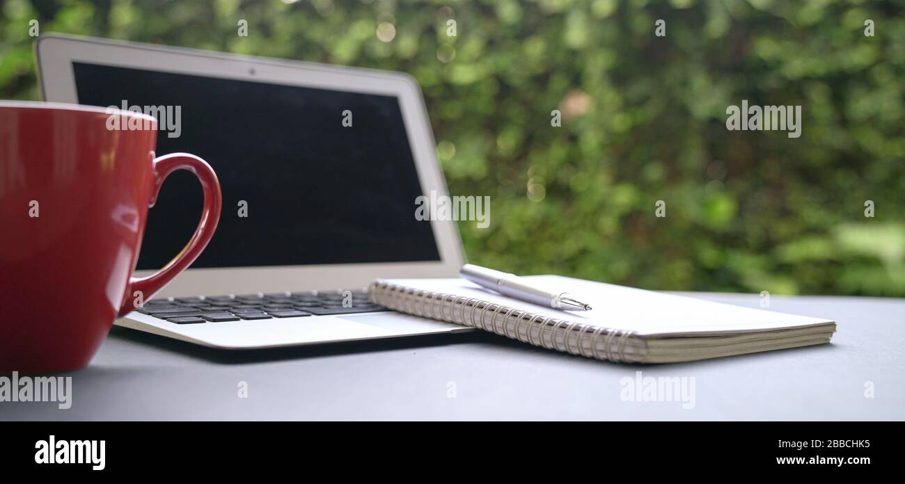 Ordinateur portable avec bloc-notes et café dans la tasse rouge avec fond vert nature. Travailler à distance ou à domicile. Espace de copie. Banque D'Images