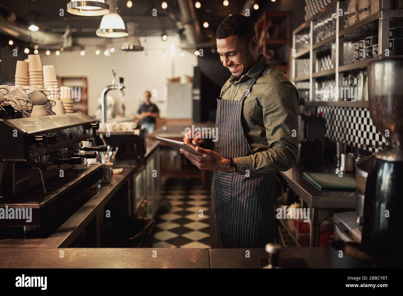 Jeune afro-américain gai propriétaire de café portant un tablier à rayures noires utilisant une tablette numérique Banque D'Images